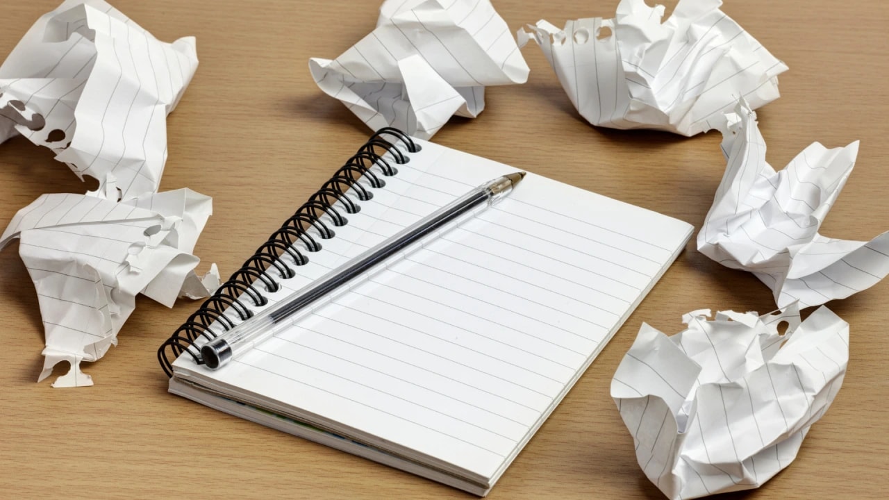 2. open spiral notebook with a pen on a wooden desk, surrounded by crumpled pieces of paper, symbolizing frustration and creative struggle.