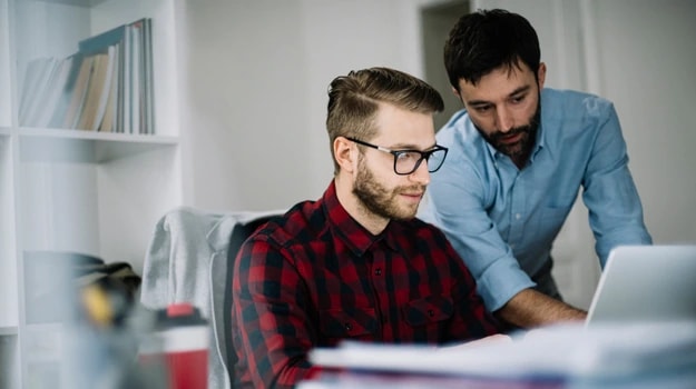 5. A man in glasses working on a laptop while another person, possibly a mentor, observes and provides guidance in an office setting.